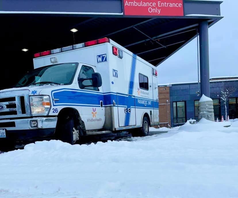 White and blue WhidbeyHealth paramedic ambulance parked in snow outside an ambulance entrance.